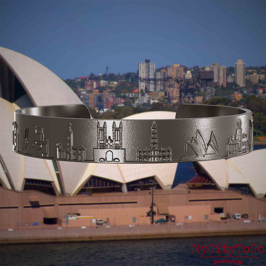Armreif aus Titan mit Reliefs bekannter Sehenswürdigkeiten der Stadt Sydney. Den Hintergrund bildet eine Ansicht der Stadt Sydney.