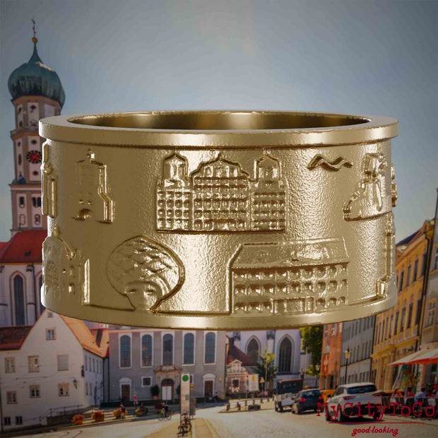 Ring aus vergoldetem Edelstahl mit Reliefs bekannter Sehenswürdigkeiten der Stadt Augsburg. Den Hintergrund bildet eine Ansicht der Stadt Augsburg.