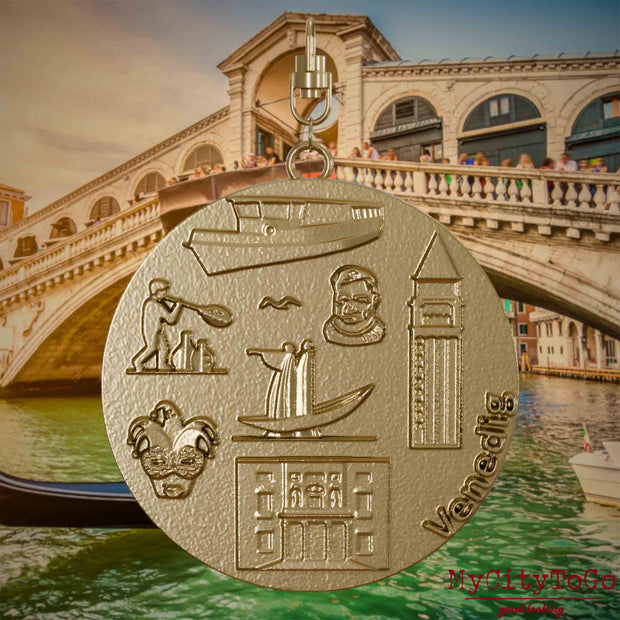 Halskette mit Drehwirbel und Rückseite eines Schmuckanhängers aus vergoldetem Edelstahl, auf dem Reliefs bekannter Sehenswürdigkeiten der Stadt Venedig abgebildet sind. Den Hintergrund bildet eine Ansicht der Stadt Venedig.
