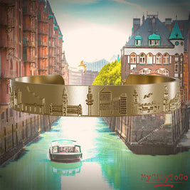 Bangle made of gold-plated stainless steel with reliefs of well-known landmarks in the city of Hamburg. The background features a view of the city of Hamburg.