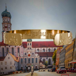 Innenseite eines vergoldeten Armreifs mit Reliefs bekannter Sehenswürdigkeiten der Stadt Augsburg und einer Gravur des Stadtnamens . Den Hintergrund bildet eine Ansicht der Stadt Augsburg.