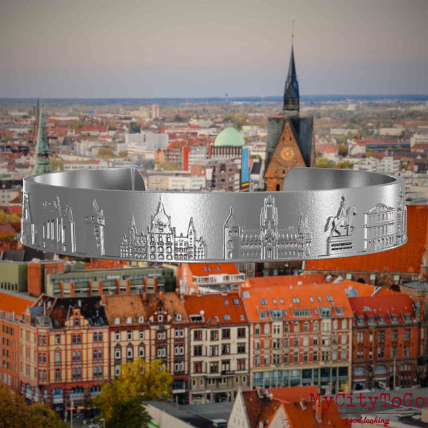 Armreif aus Edelstahl mit Reliefs bekannter Sehenswürdigkeiten der Stadt Hannover. Den Hintergrund bildet eine Ansicht der Stadt Hannover.
