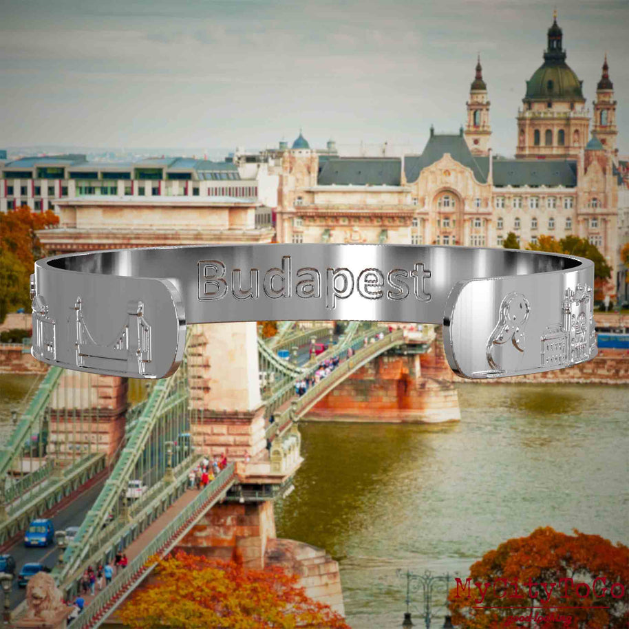 Innenseite eines silbernen Armreifs mit Reliefs bekannter Sehenswürdigkeiten der Stadt Budapest und einer Gravur des Stadtnamens. Den Hintergrund bildet eine Ansicht der Stadt Budapest.