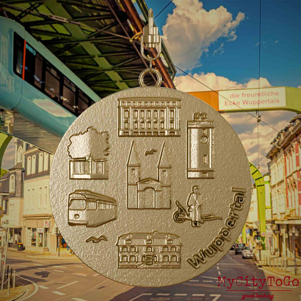 Halskette mit Drehwirbel und Rückseite eines Schmuckanhängers aus vergoldetem Edelstahl, auf dem Reliefs bekannter Sehenswürdigkeiten der Stadt Wuppertal abgebildet sind. Den Hintergrund bildet eine Ansicht der Stadt Wuppertal.
