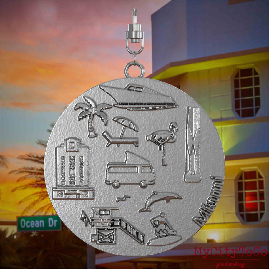 Halskette mit Drehwirbel und Rückseite eines Schmuckanhängers aus Edelstahl, auf dem Reliefs bekannter Sehenswürdigkeiten der Stadt Miami abgebildet sind. Den Hintergrund bildet eine Ansicht der Stadt Miami.
