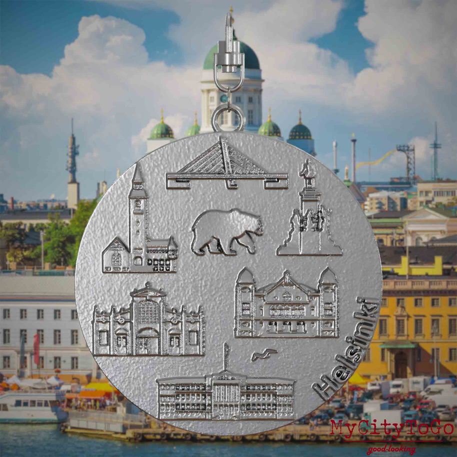 Halskette mit Drehwirbel und Rückseite eines Schmuckanhängers aus Edelstahl, auf dem Reliefs bekannter Sehenswürdigkeiten der Stadt Helsinki abgebildet sind. Den Hintergrund bildet eine Ansicht der Stadt Helsinki.
