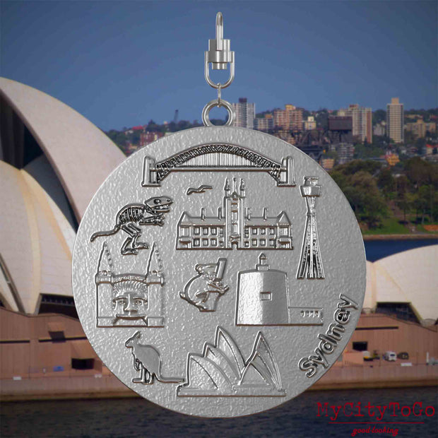 Halskette mit Drehwirbel und Vorderseite eines Schmuckanhängers aus Edelstahl, auf dem Reliefs bekannter Sehenswürdigkeiten der Stadt Sydney abgebildet sind. Den Hintergrund bildet eine Ansicht der Stadt Sydney.
