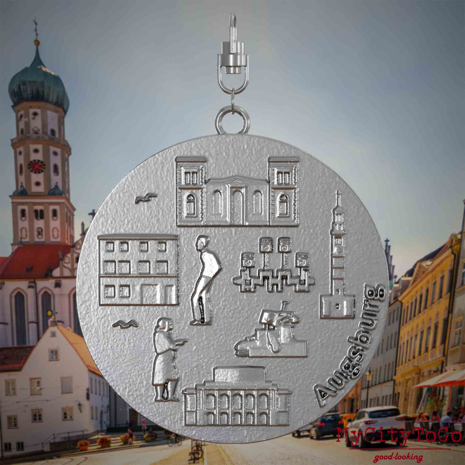 Halskette mit Drehwirbel und Vorderseite eines Schmuckanhängers aus Edelstahl, auf dem Reliefs bekannter Sehenswürdigkeiten der Stadt Augsburg abgebildet sind. Den Hintergrund bildet eine Ansicht der Stadt Augsburg .
