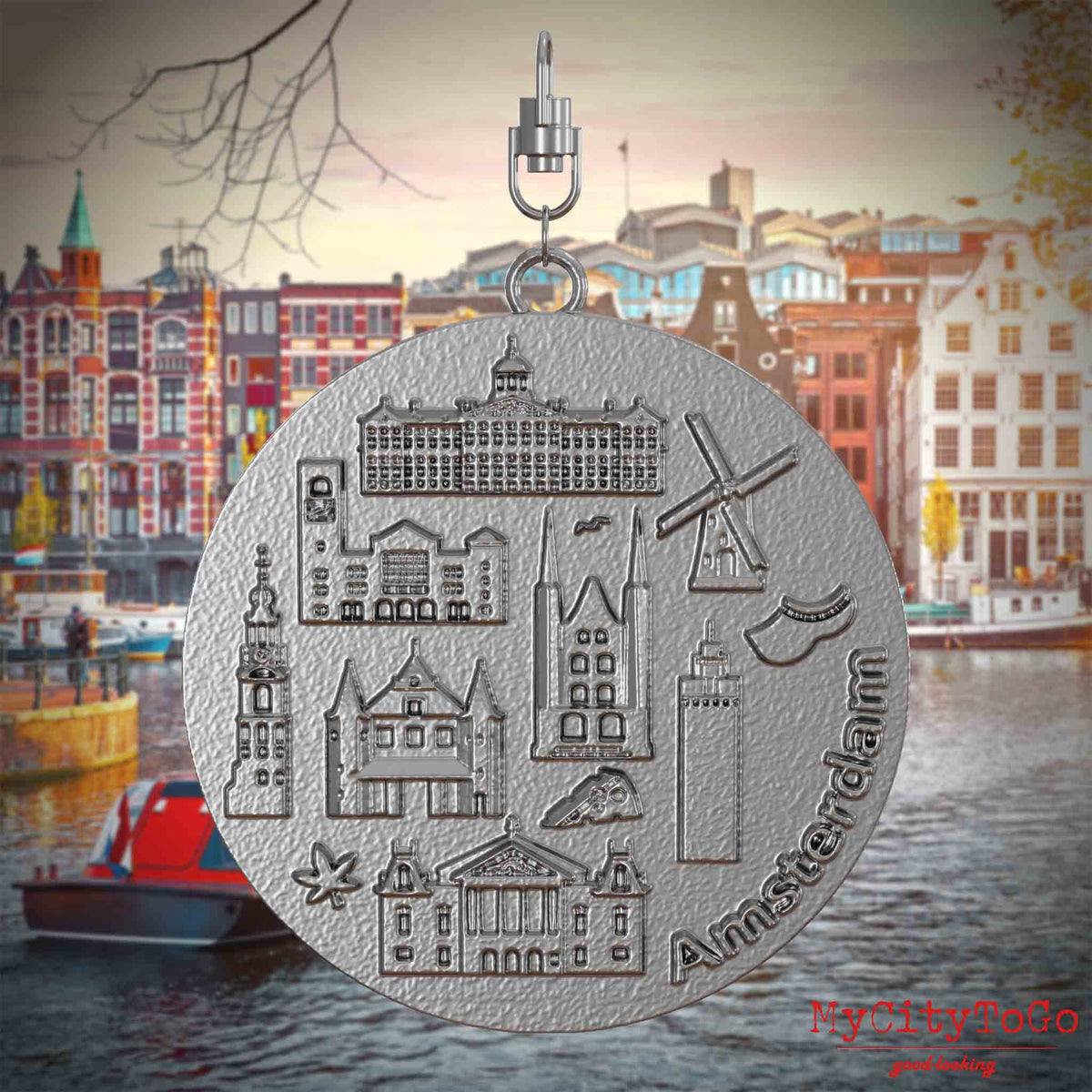 Halskette mit Drehwirbel und Rückseite eines Schmuckanhängers aus Edelstahl, auf dem Reliefs bekannter Sehenswürdigkeiten der Stadt Amsterdam abgebildet sind. Den Hintergrund bildet eine Ansicht der Stadt Amsterdam.
