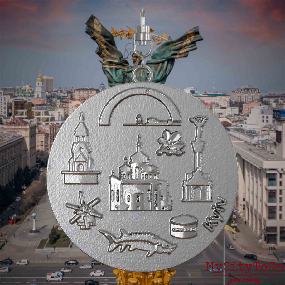 Halskette mit Drehwirbel und Rückseite eines Schmuckanhängers aus Edelstahl, auf dem Reliefs bekannter Sehenswürdigkeiten der Stadt Kyjiw abgebildet sind. Den Hintergrund bildet eine Ansicht der Stadt Kyjiw.

