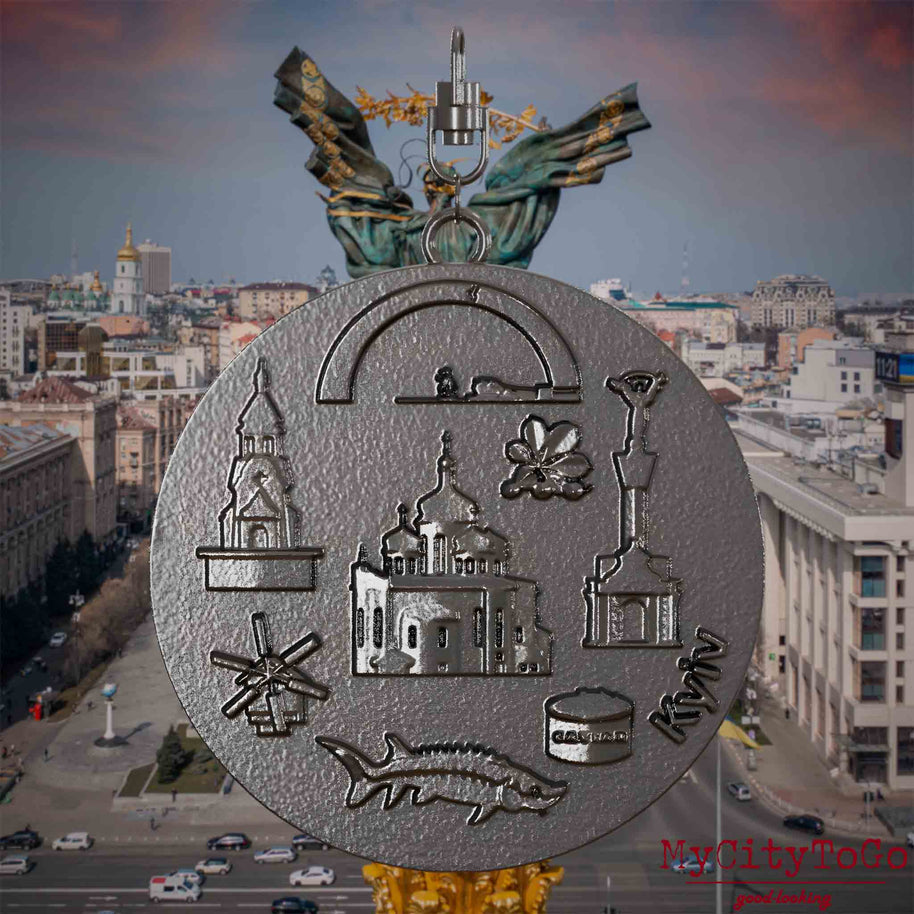 Halskette mit Drehwirbel und Rückseite eines Schmuckanhängers aus Titan, auf dem Reliefs bekannter Sehenswürdigkeiten der Stadt Kyjiw abgebildet sind. Den Hintergrund bildet eine Ansicht der Stadt Kyjiw.
