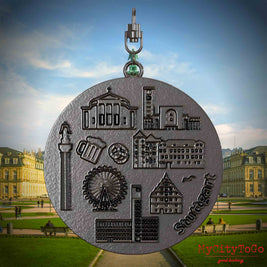 Halskette mit Drehwirbel und Rückseite eines Schmuckanhängers aus Titan, auf dem Reliefs bekannter Sehenswürdigkeiten der Stadt Stuttgart abgebildet sind. Den Hintergrund bildet eine Ansicht der Stadt Stuttgart.
