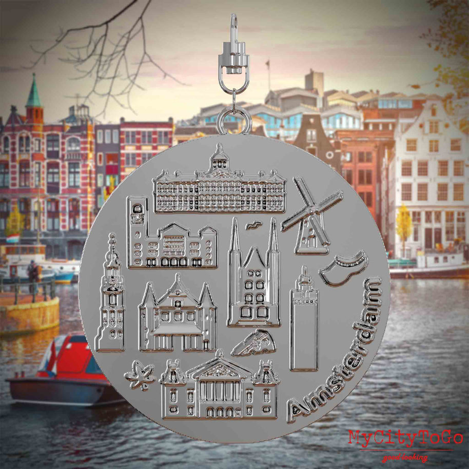 Halskette mit Drehwirbel und Rückseite eines silbernen Schmuckanhängers, auf dem Reliefs bekannter Sehenswürdigkeiten der Stadt Amsterdam abgebildet sind. Den Hintergrund bildet eine Ansicht der Stadt Amsterdam.
