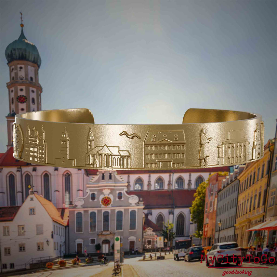 Armreif aus vergoldetem Edelstahl mit Reliefs bekannter Sehenswürdigkeiten der Stadt Augsburg. Den Hintergrund bildet eine Ansicht der Stadt Augsburg.