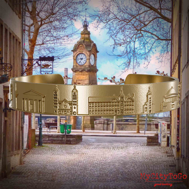 Armreif aus vergoldetem Edelstahl mit Reliefs bekannter Sehenswürdigkeiten der Stadt Duesseldorf. Den Hintergrund bildet eine Ansicht der Stadt Duesseldorf.