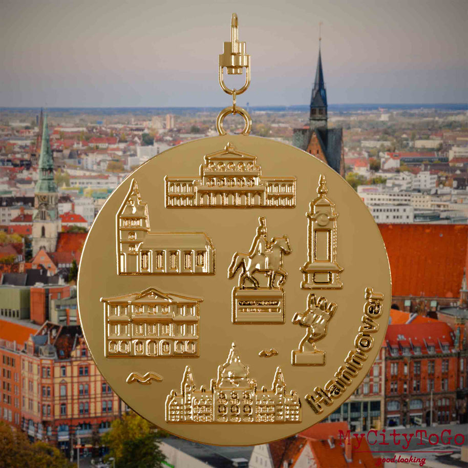 Halskette mit Drehwirbel und Rückseite eines vergoldeten Schmuckanhängers, auf dem Reliefs bekannter Sehenswürdigkeiten der Stadt Hannover abgebildet sind. Den Hintergrund bildet eine Ansicht der Stadt Hannover .
