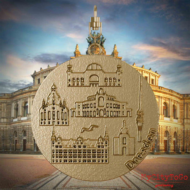 Halskette mit Drehwirbel und Rückseite eines Schmuckanhängers aus vergoldetem Edelstahl, auf dem Reliefs bekannter Sehenswürdigkeiten der Stadt Dresden abgebildet sind. Den Hintergrund bildet eine Ansicht der Stadt Dresden.