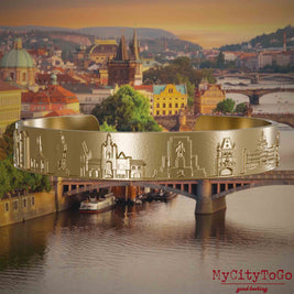 Armreif aus vergoldetem Edelstahl mit Reliefs bekannter Sehenswürdigkeiten der Stadt Prag. Den Hintergrund bildet eine Ansicht der Stadt Prag.