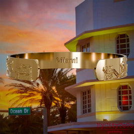 Innenseite eines vergoldeten Armreifs mit Reliefs bekannter Sehenswürdigkeiten der Stadt Miami und einer Gravur des Stadtnamens. Den Hintergrund bildet eine Ansicht der Stadt Miami.