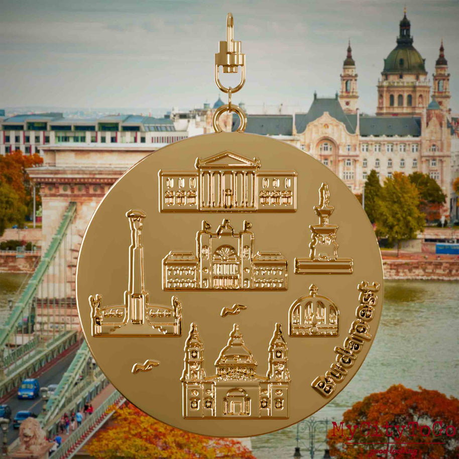 Halskette mit Drehwirbel und Rückseite eines vergoldeten Schmuckanhängers, auf dem Reliefs bekannter Sehenswürdigkeiten der Stadt Budapest abgebildet sind. Den Hintergrund bildet eine Ansicht der Stadt Budapest.
