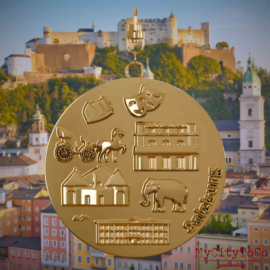 Halskette mit Drehwirbel und Rückseite eines vergoldeten Schmuckanhängers, auf dem Reliefs bekannter Sehenswürdigkeiten der Stadt Salzburg abgebildet sind. Den Hintergrund bildet eine Ansicht der Stadt Salzburg.
