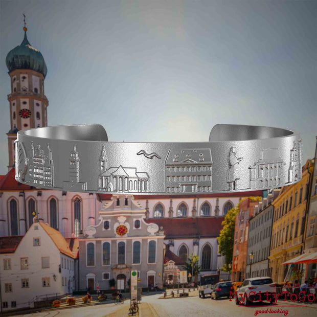 Armreif aus Edelstahl mit Reliefs bekannter Sehenswürdigkeiten der Stadt Augsburg. Den Hintergrund bildet eine Ansicht der Stadt Augsburg.