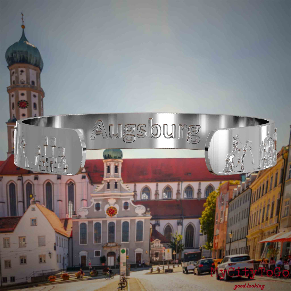 Innenseite eines silbernen Armreifs mit Reliefs bekannter Sehenswürdigkeiten der Stadt Augsburg und einer Gravur des Stadtnamens . Den Hintergrund bildet eine Ansicht der Stadt Augsburg.