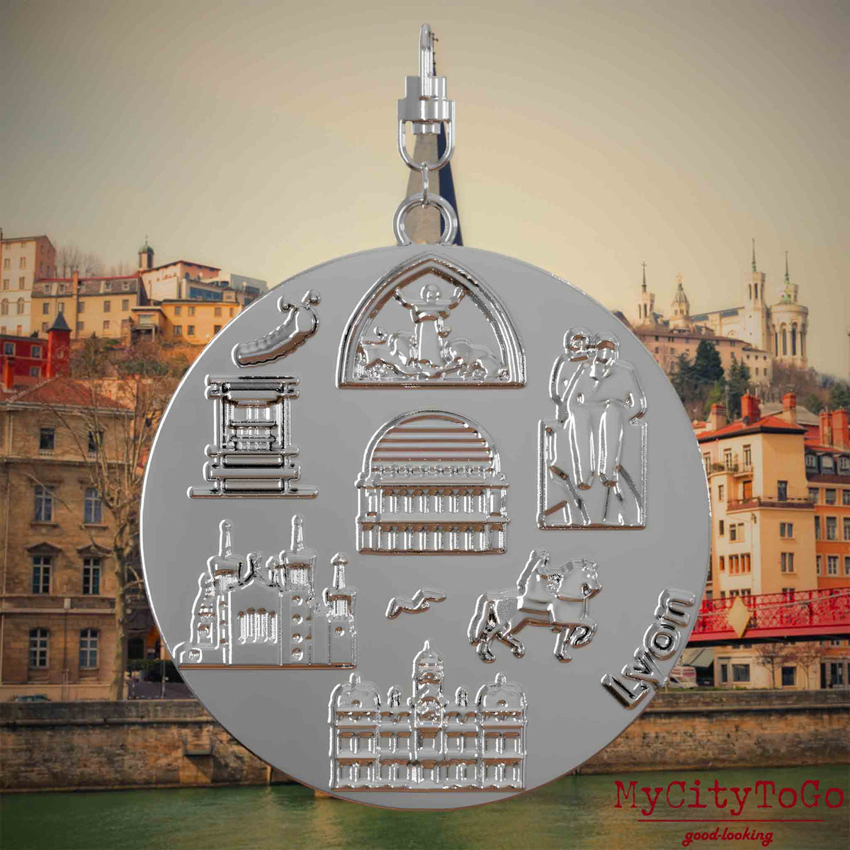 Halskette mit Drehwirbel und Rückseite eines silbernen Schmuckanhängers, auf dem Reliefs bekannter Sehenswürdigkeiten der Stadt Lyon abgebildet sind. Den Hintergrund bildet eine Ansicht der Stadt Lyon.
