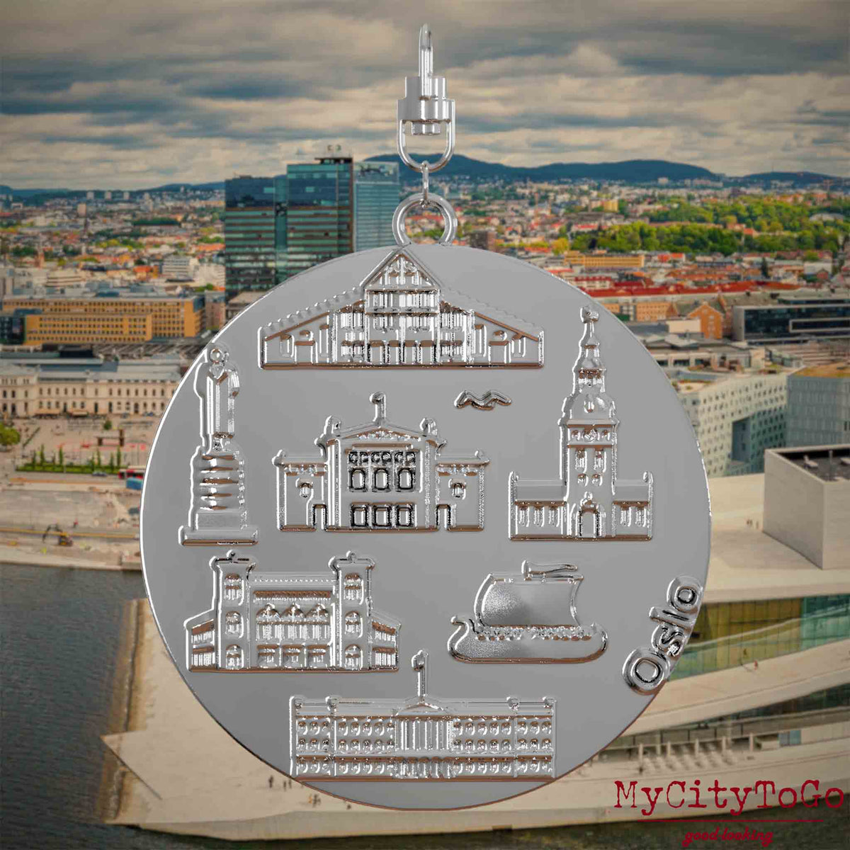 Halskette mit Drehwirbel und Rückseite eines silbernen Schmuckanhängers, auf dem Reliefs bekannter Sehenswürdigkeiten der Stadt Oslo abgebildet sind. Den Hintergrund bildet eine Ansicht der Stadt Oslo.
