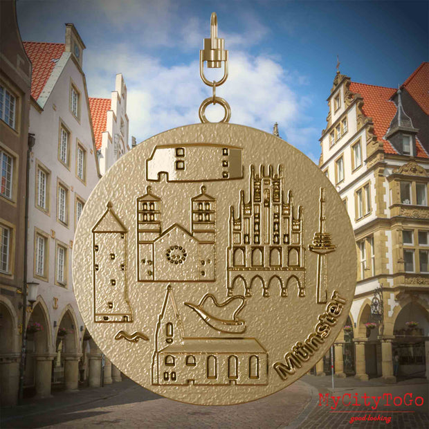 Halskette mit Drehwirbel und Vorderseite eines Schmuckanhängers aus vergoldetem Edelstahl, auf dem Reliefs bekannter Sehenswürdigkeiten der Stadt Muenster abgebildet sind. Den Hintergrund bildet eine Ansicht der Stadt Muenster.