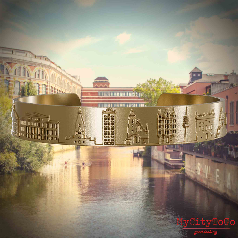 Armreif aus vergoldetem Edelstahl mit Reliefs bekannter Sehenswürdigkeiten der Stad Leipzig. Den Hintergrund bildet eine Ansicht der Stadt Leipzig.