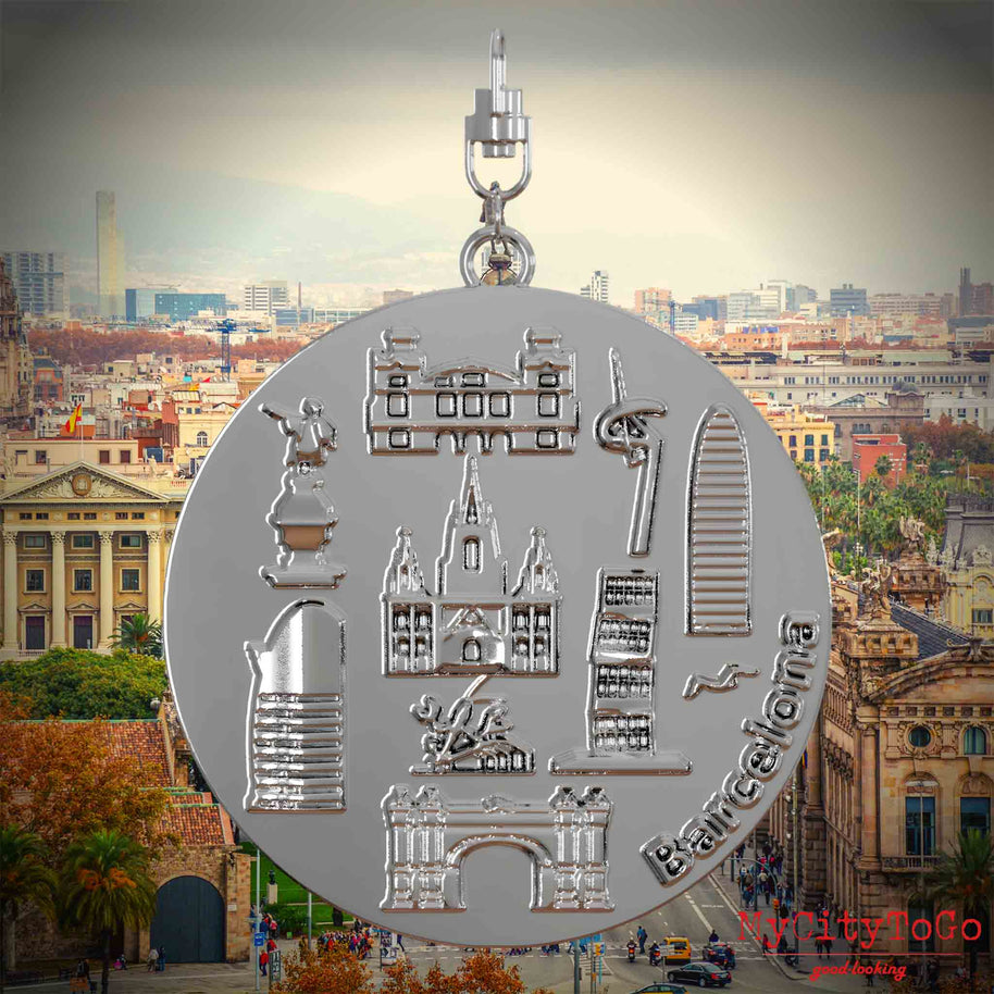 Halskette mit Drehwirbel und Vorderseite eines silbernen Schmuckanhängers, auf dem Reliefs bekannter Sehenswürdigkeiten der Stadt Barcelona abgebildet sind. Den Hintergrund bildet eine Ansicht der Stadt Barcelona.
