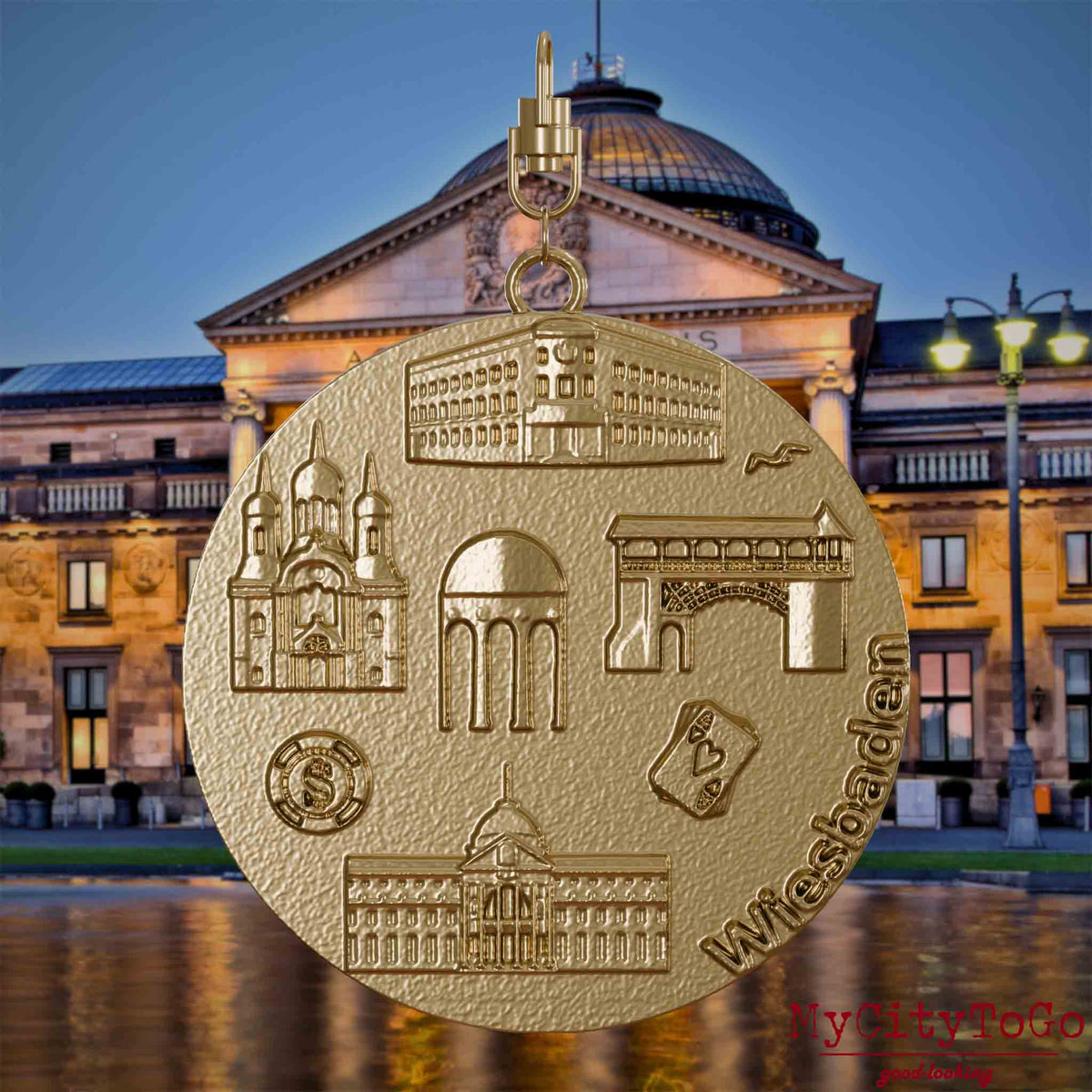Halskette mit Drehwirbel und Vorderseite eines Schmuckanhängers aus vergoldetem Edelstahl, auf dem Reliefs bekannter Sehenswürdigkeiten der Stadt Wiesbaden abgebildet sind. Den Hintergrund bildet eine Ansicht der Stadt Wiesbaden.
