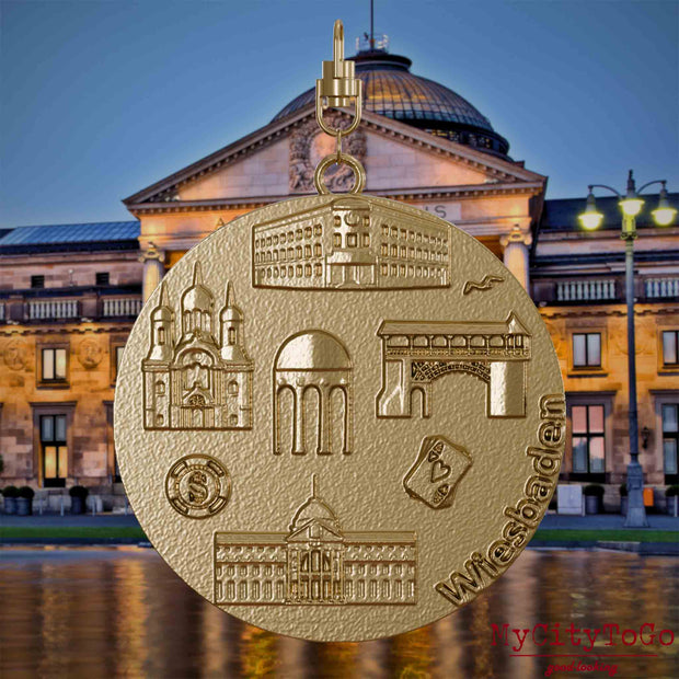 Halskette mit Drehwirbel und Vorderseite eines Schmuckanhängers aus vergoldetem Edelstahl, auf dem Reliefs bekannter Sehenswürdigkeiten der Stadt Wiesbaden abgebildet sind. Den Hintergrund bildet eine Ansicht der Stadt Wiesbaden.
