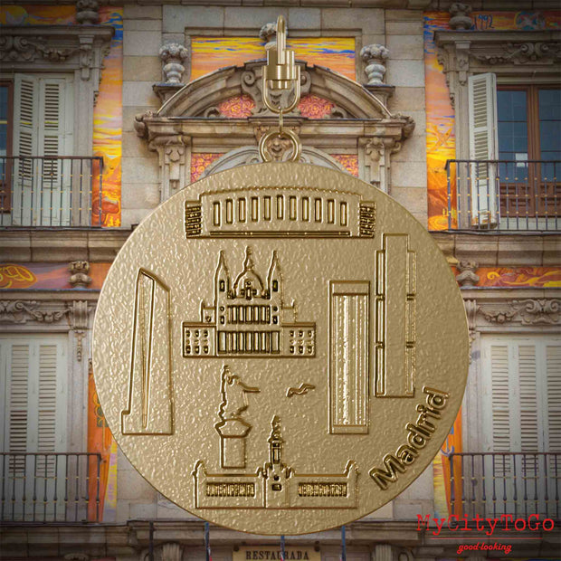 Halskette mit Drehwirbel und Rückseite eines Schmuckanhängers aus vergoldetem Edelstahl, auf dem Reliefs bekannter Sehenswürdigkeiten der Stadt Madrid abgebildet sind. Den Hintergrund bildet eine Ansicht der Stadt Madrid.
