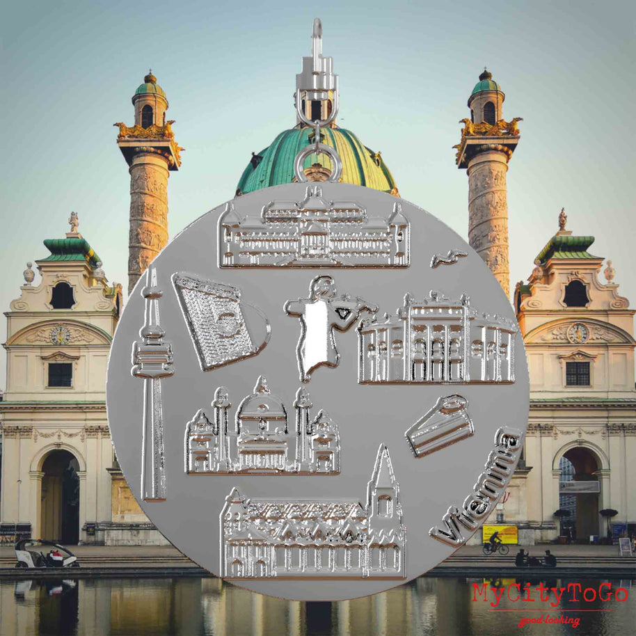 Halskette mit Drehwirbel und Rückseite eines silbernen Schmuckanhängers, auf dem Reliefs bekannter Sehenswürdigkeiten der Stadt Wien abgebildet sind. Den Hintergrund bildet eine Ansicht der Stadt Wien.
