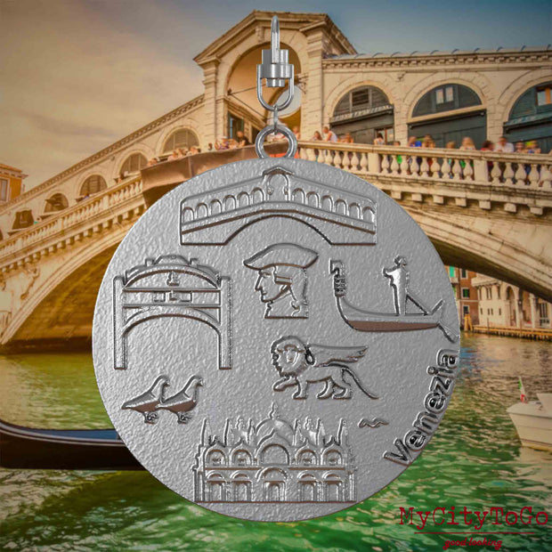 Halskette mit Drehwirbel und Vorderseite eines Schmuckanhängers aus Edelstahl, auf dem Reliefs bekannter Sehenswürdigkeiten der Stadt Venedig abgebildet sind. Den Hintergrund bildet eine Ansicht der Stadt Venedig.
