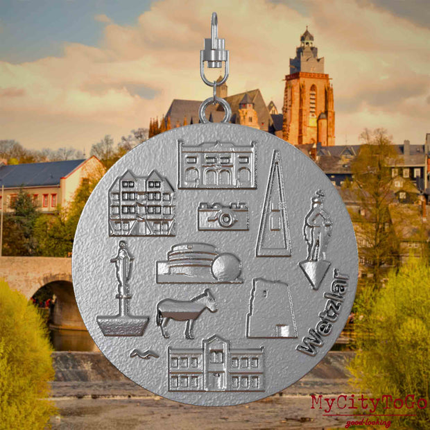 Halskette mit Drehwirbel und Vorderseite eines Schmuckanhängers aus Edelstahl, auf dem Reliefs bekannter Sehenswürdigkeiten der Stadt Wetzlar abgebildet sind. Den Hintergrund bildet eine Ansicht der Stadt Wetzlar.

