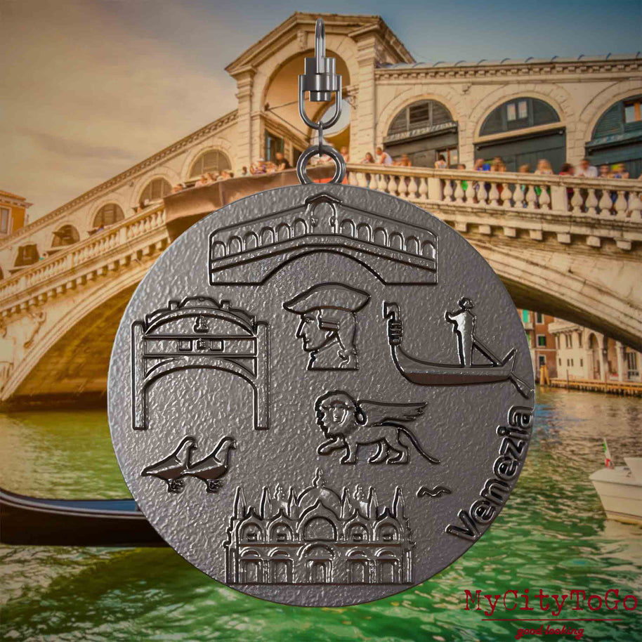 Halskette mit Drehwirbel und Vorderseite eines Schmuckanhängers aus Titan, auf dem Reliefs bekannter Sehenswürdigkeiten der Stadt Venedig abgebildet sind. Den Hintergrund bildet eine Ansicht der Stadt Venedig.
