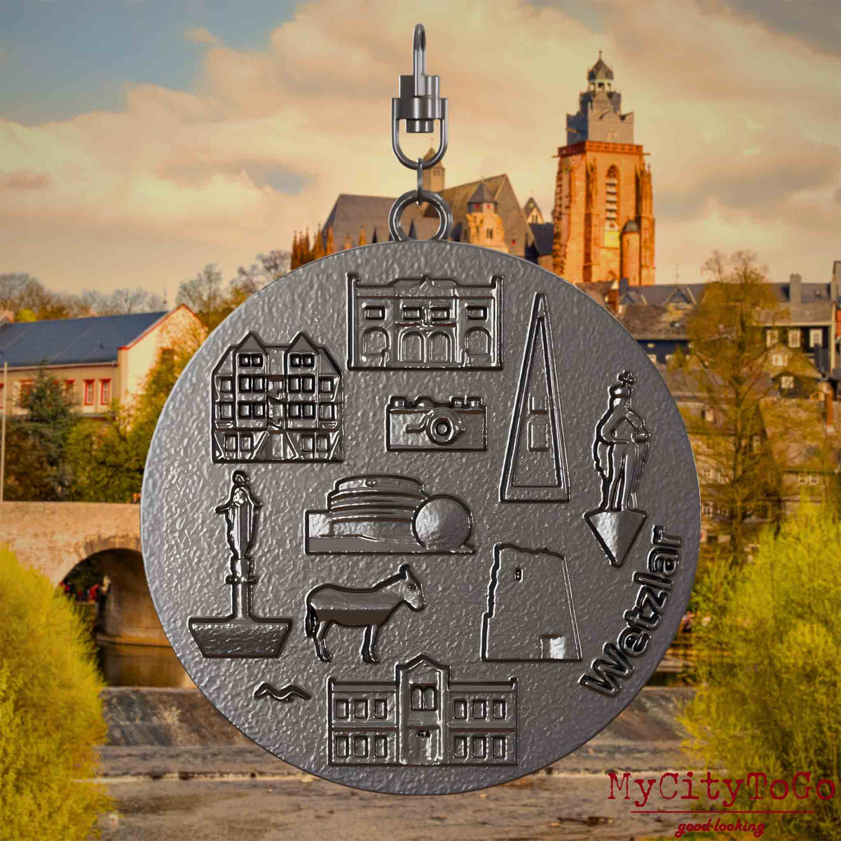 Halskette mit Drehwirbel und Vorderseite eines Schmuckanhängers aus Titan, auf dem Reliefs bekannter Sehenswürdigkeiten der Stadt Wetzlar abgebildet sind. Den Hintergrund bildet eine Ansicht der Stadt Wetzlar.
