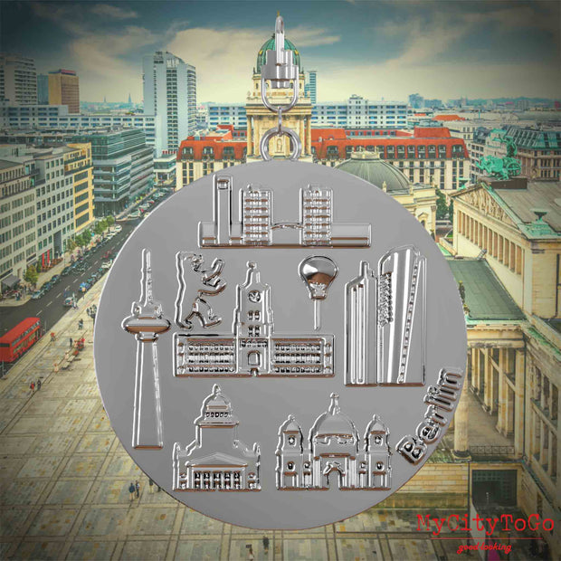 Halskette mit Drehwirbel und Vorderseite eines silbernen Schmuckanhängers, auf dem Reliefs bekannter Sehenswürdigkeiten der Stadt Berlin abgebildet sind. Den Hintergrund bildet eine Ansicht der Stadt Berlin.