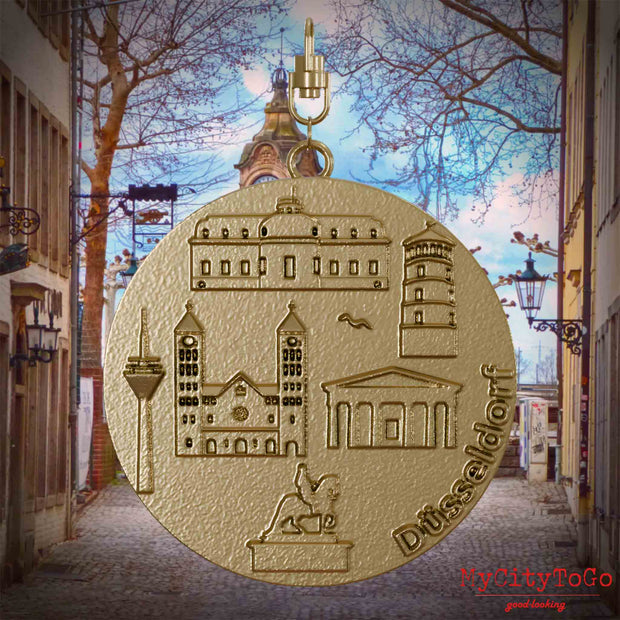 Halskette mit Drehwirbel und Vorderseite eines Schmuckanhängers aus vergoldetem Edelstahl, auf dem Reliefs bekannter Sehenswürdigkeiten der Stadt Duesseldorf abgebildet sind. Den Hintergrund bildet eine Ansicht der Stadt Duesseldorf.
