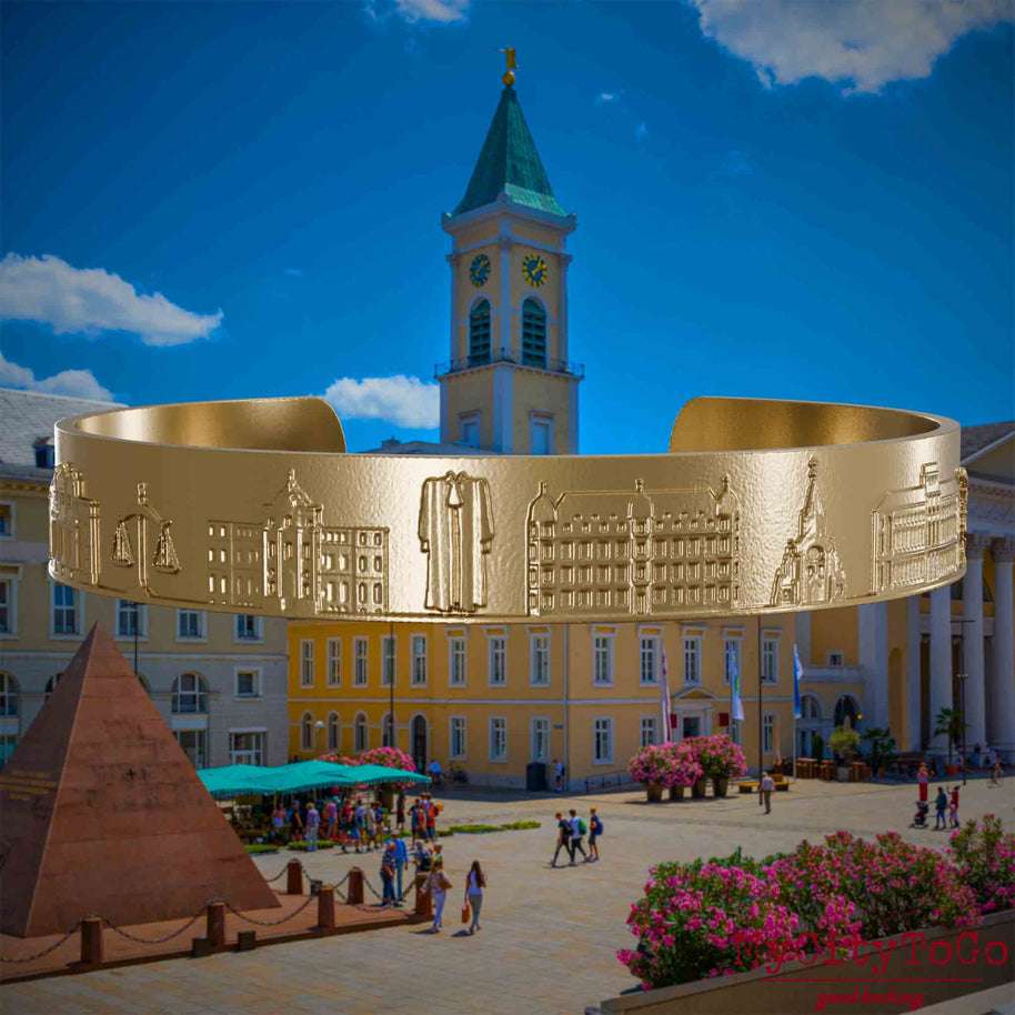 Armreif aus vergoldetem Edelstahl mit Reliefs bekannter Sehenswürdigkeiten der Stadt Karlsruhe. Den Hintergrund bildet eine Ansicht der Stadt Karlsruhe.