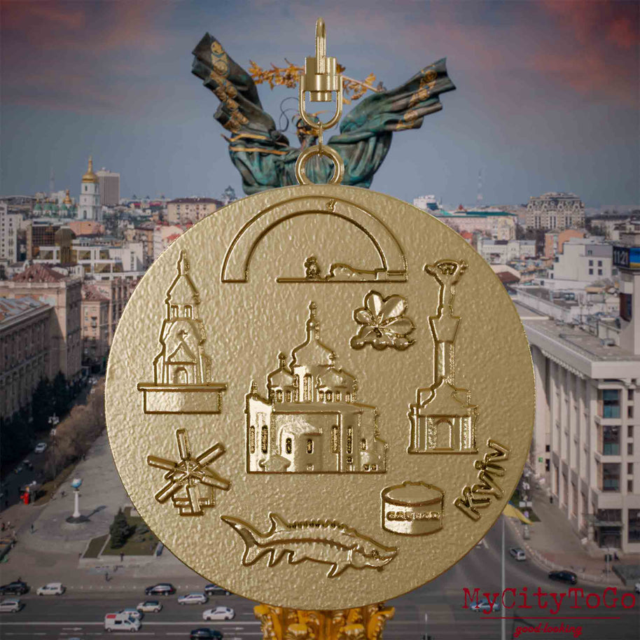 Halskette mit Drehwirbel und Rückseite eines Schmuckanhängers aus vergoldetem Edelstahl, auf dem Reliefs bekannter Sehenswürdigkeiten der Stadt Kyjiw abgebildet sind. Den Hintergrund bildet eine Ansicht der Stadt Kyjiw.
