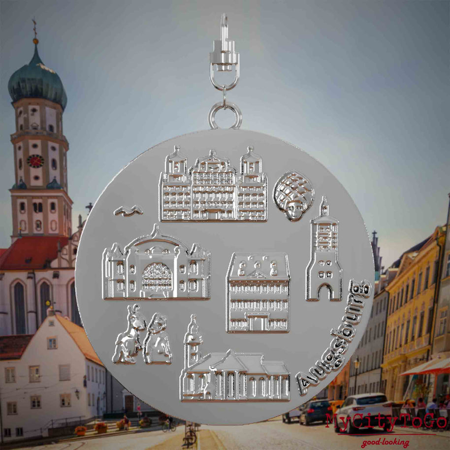 Halskette mit Drehwirbel und Rückseite eines silbernen Schmuckanhängers, auf dem Reliefs bekannter Sehenswürdigkeiten der Stadt Augsburg abgebildet sind. Den Hintergrund bildet eine Ansicht der Stadt Augsburg .
