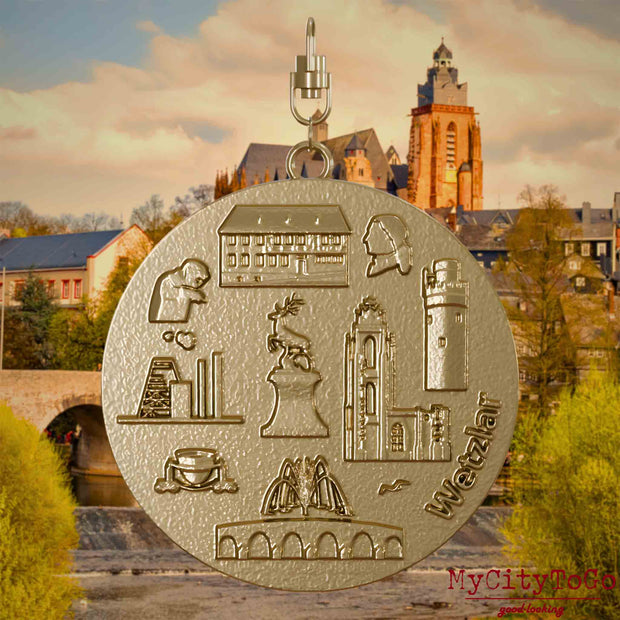 Halskette mit Drehwirbel und Rückseite eines Schmuckanhängers aus vergoldetem Edelstahl, auf dem Reliefs bekannter Sehenswürdigkeiten der Stadt Wetzlar abgebildet sind. Den Hintergrund bildet eine Ansicht der Stadt Wetzlar.
