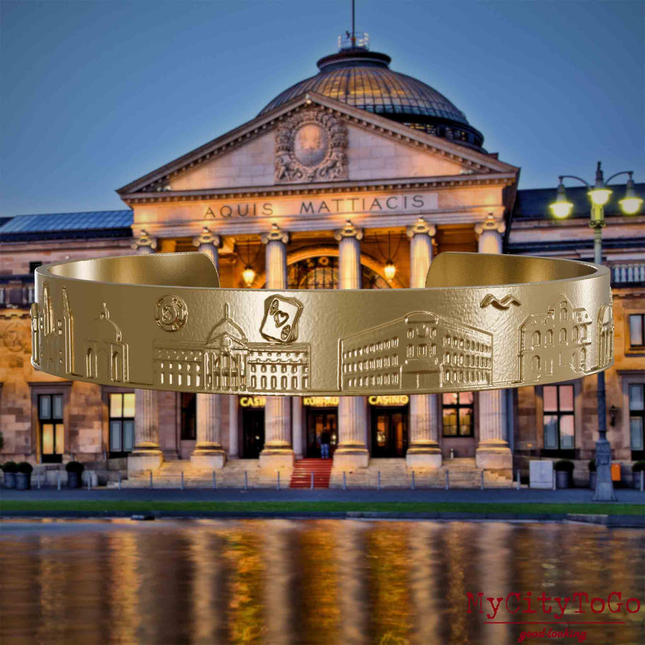 Bangle made of gold-plated stainless steel with reliefs of well-known landmarks of the city of Wiesbaden. The background features a view of the city of Wiesbaden.
