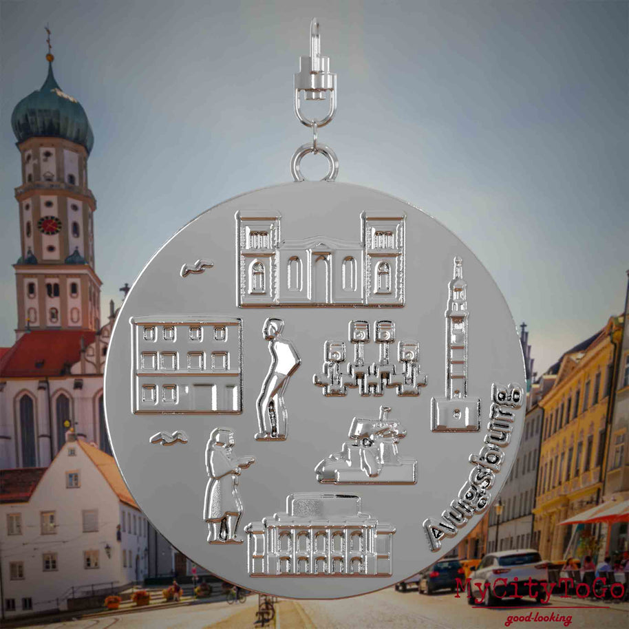Halskette mit Drehwirbel und Vorderseite eines silbernen Schmuckanhängers, auf dem Reliefs bekannter Sehenswürdigkeiten der Stadt Augsburg abgebildet sind. Den Hintergrund bildet eine Ansicht der Stadt Augsburg .

