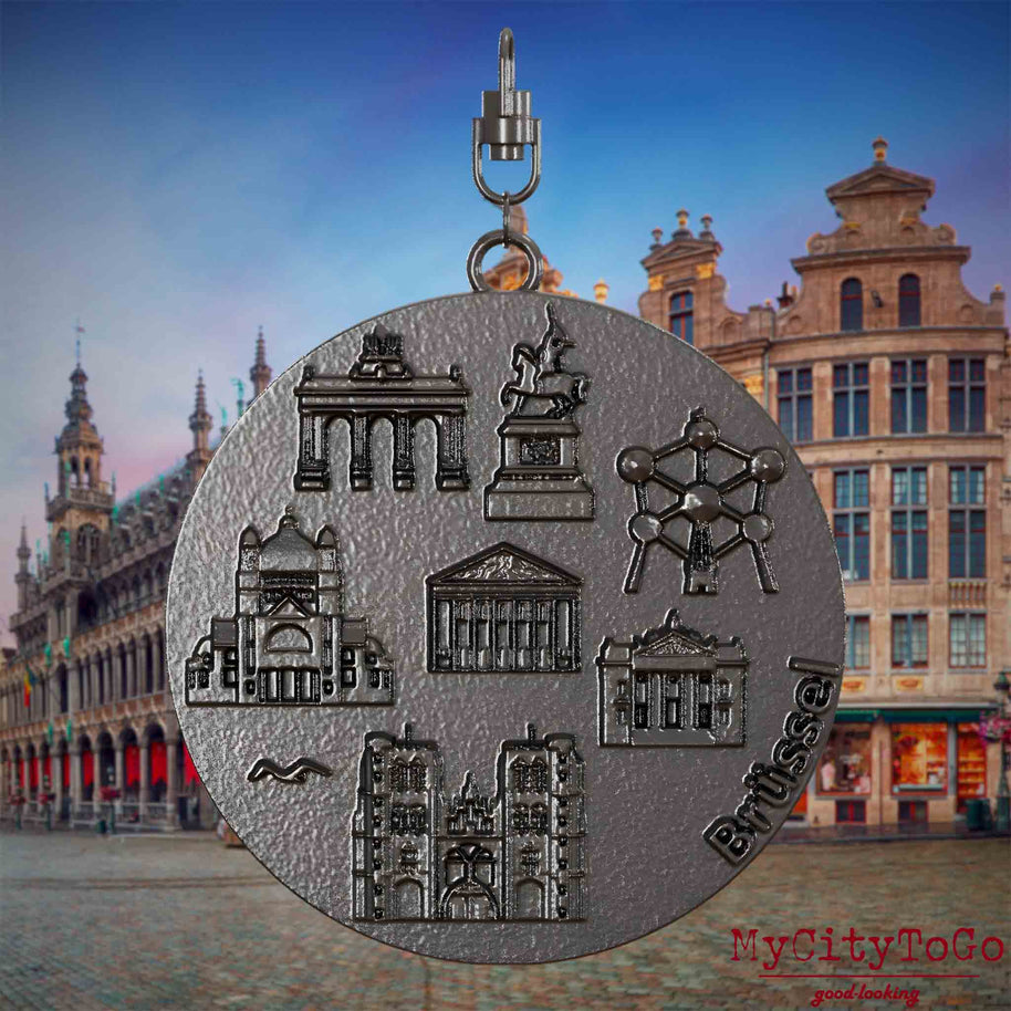 Halskette mit Drehwirbel und Rückseite eines Schmuckanhängers aus Titan, auf dem Reliefs bekannter Sehenswürdigkeiten der Stadt Bruessel abgebildet sind. Den Hintergrund bildet eine Ansicht der Stadt Bruessel.
