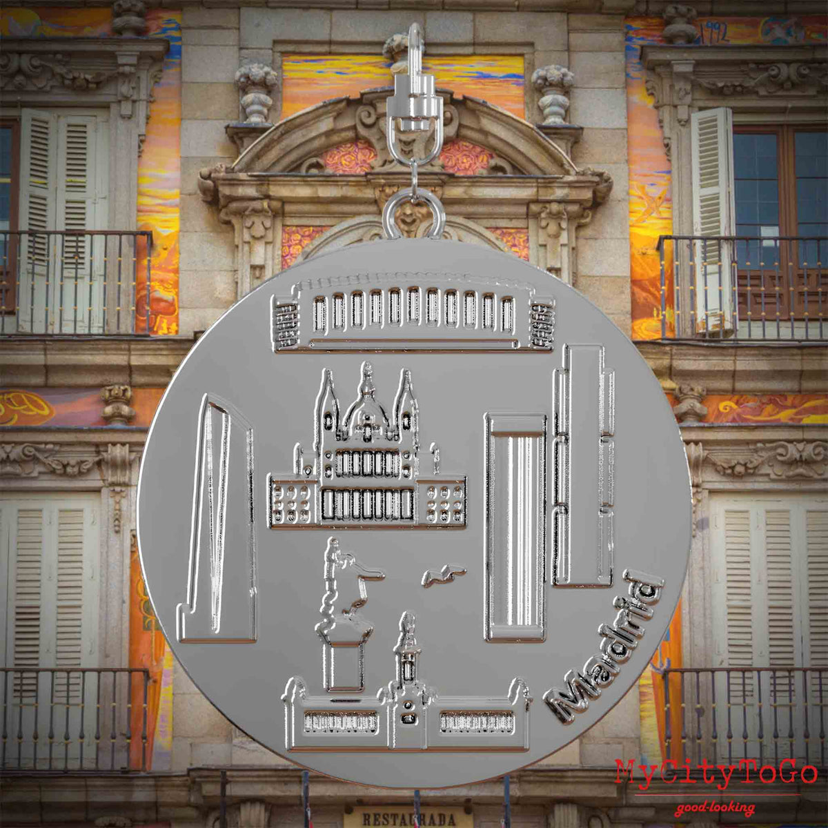 Halskette mit Drehwirbel und Rückseite eines silbernen Schmuckanhängers, auf dem Reliefs bekannter Sehenswürdigkeiten der Stadt Madrid abgebildet sind. Den Hintergrund bildet eine Ansicht der Stadt Madrid.
